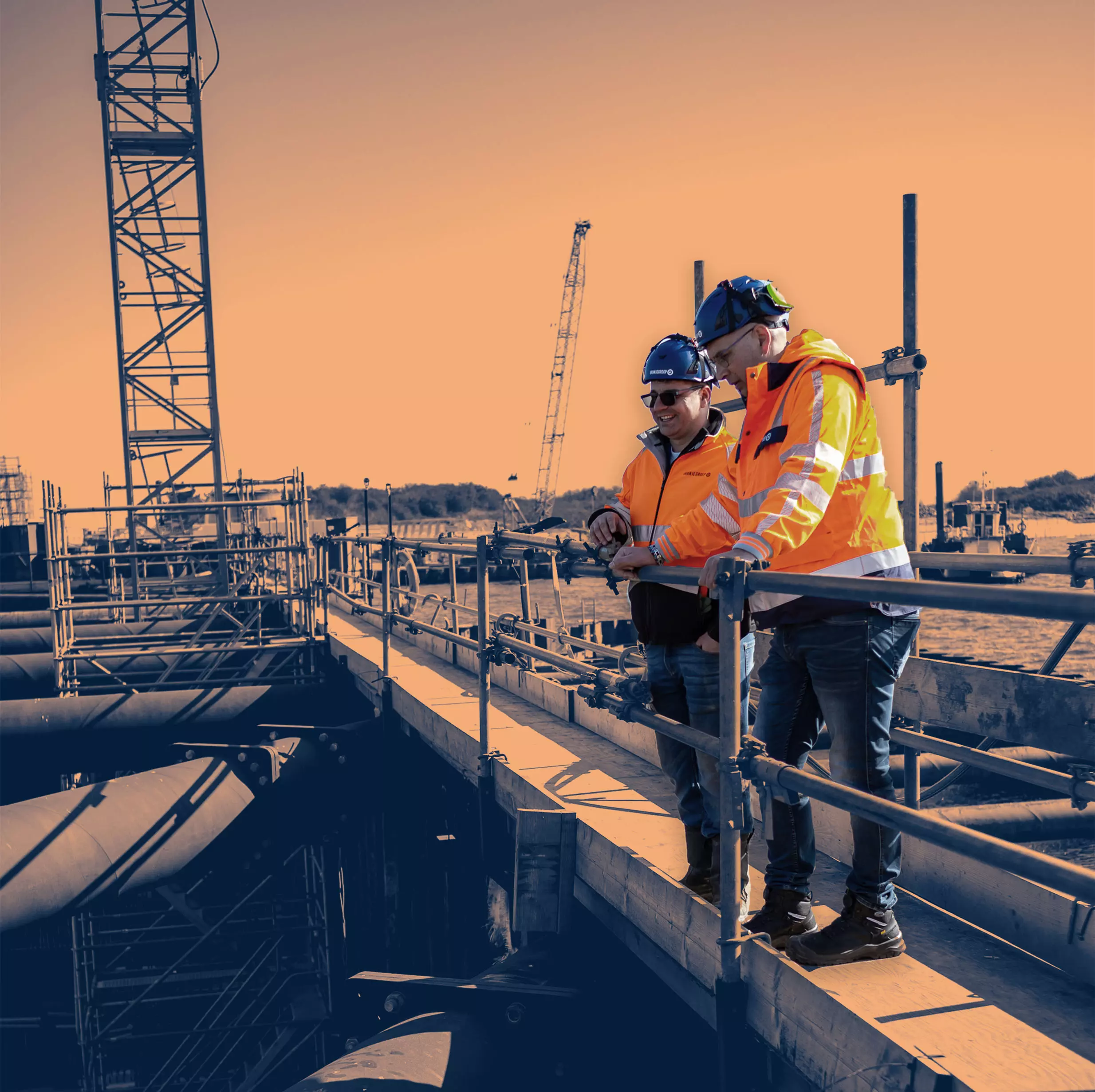 Shared Success Begins on Site
Two construction supervisors in safety gear discussing project progress on-site, standing on a platform above large industrial pipes.